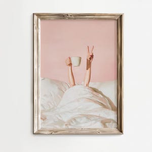 May include: A framed print depicts a person in bed, holding a white mug and making a peace sign. The artwork has a pink background and a white comforter. The frame is a light brown color.