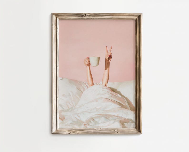 May include: A framed print depicts a person in bed, holding a white mug and making a peace sign. The artwork has a pink background and a white comforter. The frame is a light brown color.