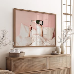 May include: A framed print depicts a person pouring champagne into a glass while in bed. The artwork has a pink background and a brown frame. The image is displayed above a wooden cabinet with decorative vases and a woven basket.