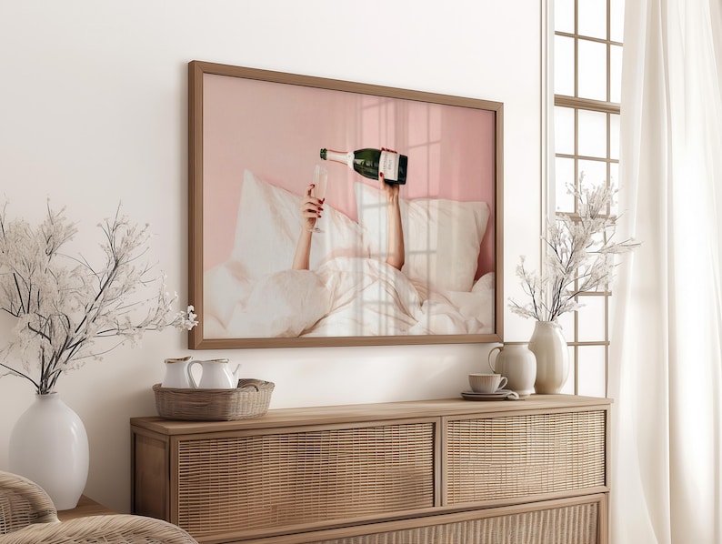 May include: A framed print depicts a person pouring champagne into a glass while in bed. The artwork has a pink background and a brown frame. The image is displayed above a wooden cabinet with decorative vases and a woven basket.