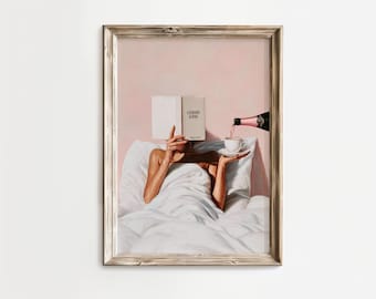 May include: A framed print depicts a person in bed reading a book titled "FASHION ICONS" while champagne is poured into a teacup. The artwork has a soft, pink and white color palette, with a vintage aesthetic.