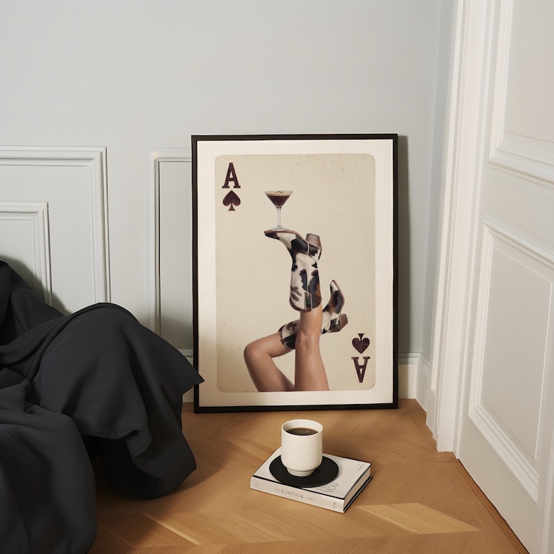 Pode incluir: Impress&atilde;o art&iacute;stica emoldurada com um copo de cocktail equilibrado em pernas com botas com estampa de vaca, estilizado como um &Aacute;s de Espadas. Uma ch&aacute;vena de caf&eacute; branca repousa sobre um livro.