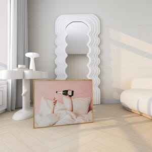 May include: A framed print with a gold border depicts a person pouring champagne into a glass. The image is set against a pink background with white pillows and bedding. A large white mirror with a wavy design is in the background.