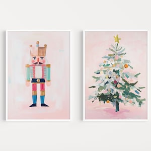 Puede incluir: Dos impresiones artísticas enmarcadas. Una presenta un colorido soldado cascanueces con corona, y la otra un árbol de Navidad con adornos y una estrella. Ambas están pintadas sobre un fondo rosa.