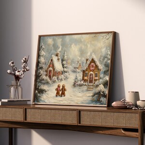 May include: A framed painting depicts a winter scene with snow-covered gingerbread houses and trees. Two gingerbread people stand in the foreground. The artwork is in a wooden frame and evokes a cozy, festive atmosphere.