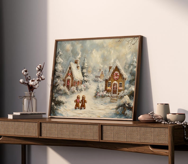 May include: A framed painting depicts a winter scene with snow-covered gingerbread houses and trees. Two gingerbread people stand in the foreground. The artwork is in a wooden frame and evokes a cozy, festive atmosphere.