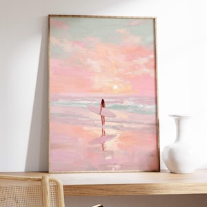 May include: A framed art print depicts a serene beach scene at sunset. A person stands in the water holding a surfboard. The sky is painted with soft pinks and blues, reflecting on the wet sand. The artwork is framed in a light wood.