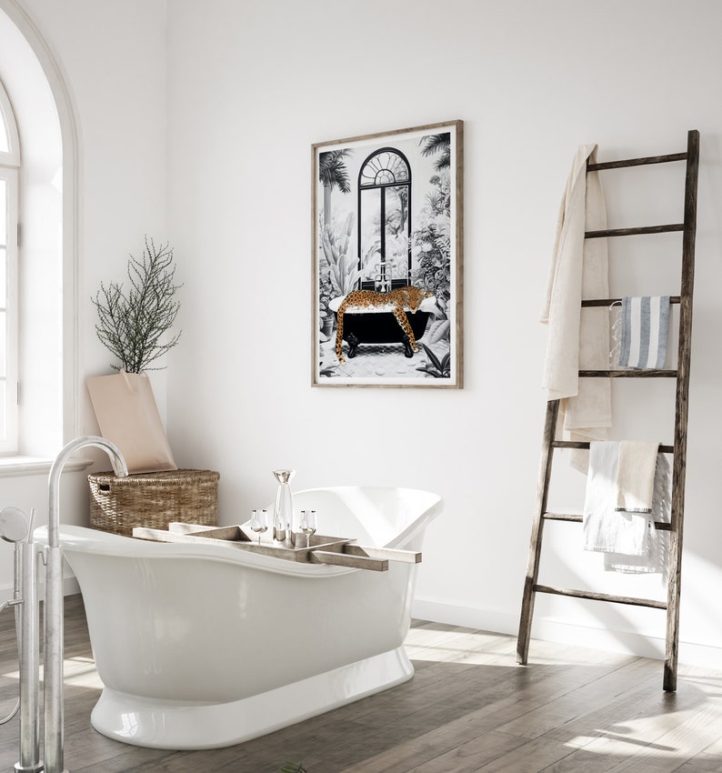 May include: A framed wall art print featuring a black and white illustration of a leopard resting in a clawfoot bathtub surrounded by tropical plants, displayed in a bathroom with a white freestanding tub and a wooden towel ladder.