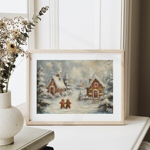 May include: A framed print of a winter scene featuring two gingerbread houses covered in snow. Two gingerbread people walk on a snowy path. The artwork is in a light wooden frame, set against a white background.
