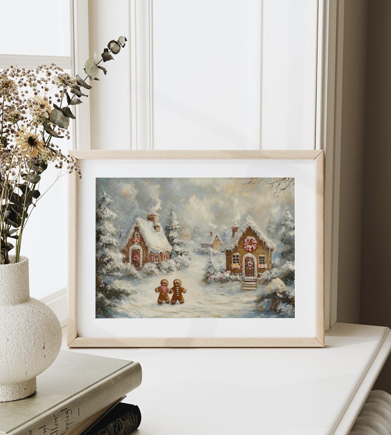 May include: A framed print of a winter scene featuring two gingerbread houses covered in snow. Two gingerbread people walk on a snowy path. The artwork is in a light wooden frame, set against a white background.