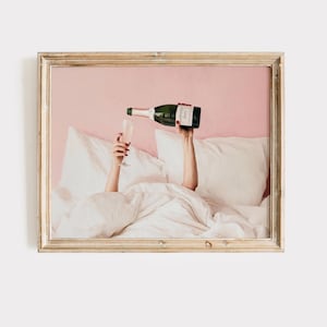 May include: A framed print shows a person pouring champagne into a glass. The person is in bed, surrounded by white pillows and a comforter. The champagne bottle is green and the background is pink.
