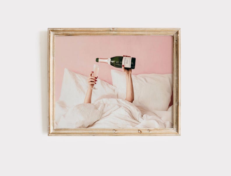 May include: A framed print shows a person pouring champagne into a glass. The person is in bed, surrounded by white pillows and a comforter. The champagne bottle is green and the background is pink.