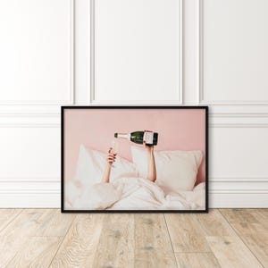 May include: A framed print shows a person pouring champagne into a glass. The person is in bed, with white pillows and a duvet. The champagne bottle is green with a white label. The background is pink.