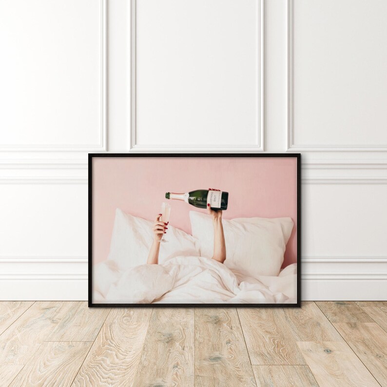 May include: A framed print shows a person pouring champagne into a glass. The person is in bed, with white pillows and a duvet. The champagne bottle is green with a white label. The background is pink.