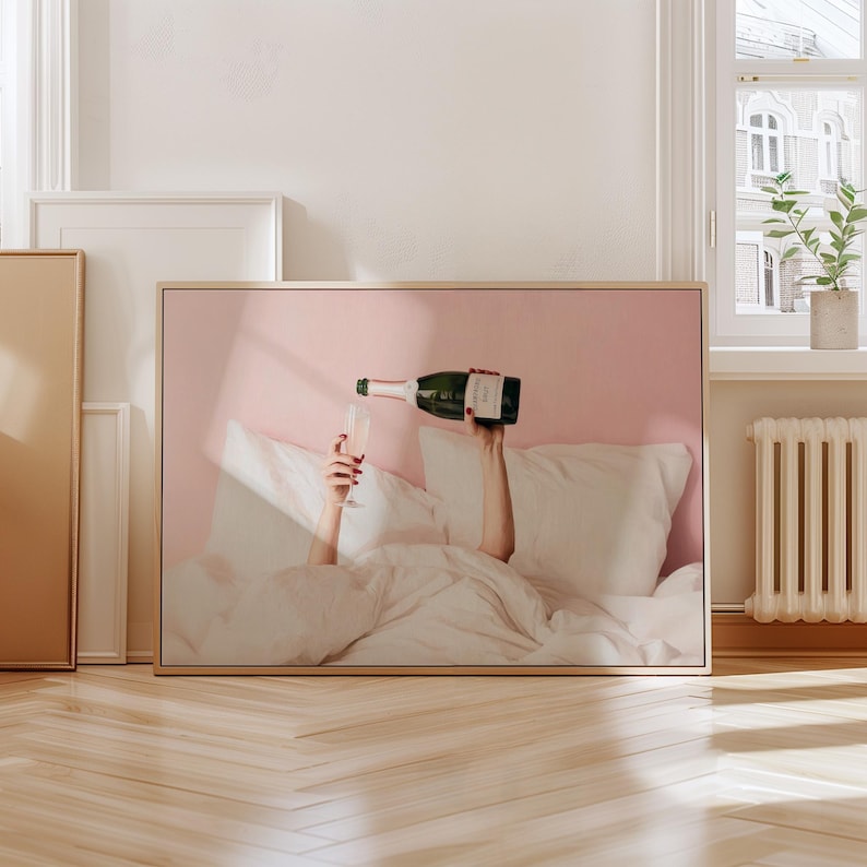May include: A framed print depicts a person in bed pouring champagne into a glass. The image has a pink background and a light wooden frame. The person's hands and the champagne bottle are visible.