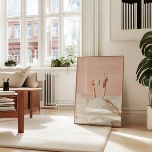 May include: A framed print featuring a person in bed holding a coffee cup and making a peace sign. The artwork has a pink background and a copper-colored frame. The room includes a sofa, coffee table, and a large potted plant.
