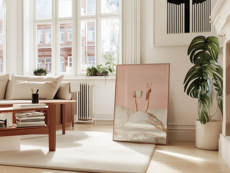 May include: A framed print featuring a person in bed holding a coffee cup and making a peace sign. The artwork has a pink background and a copper-colored frame. The room includes a sofa, coffee table, and a large potted plant.