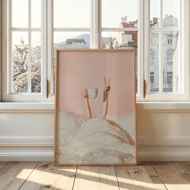 May include: A framed art print featuring a person's arms reaching up from under a white blanket. One hand holds a white mug, the other makes a peace sign. The background is a soft pink, with a wooden frame.