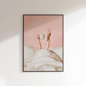 May include: Framed art print featuring a person's arms reaching out from under a white blanket. One hand holds a white mug, the other makes a peace sign. The background is a solid pink color.