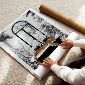 May include: A square art print featuring a black and white illustration of a large arched window surrounded by lush tropical foliage and palm leaves. A realistic, colored leopard is depicted lying across the bottom section of the window frame.
