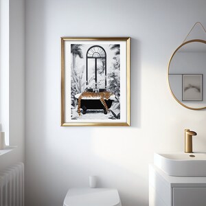 May include: A framed wall art print featuring a black and white illustration of a leopard resting in a clawfoot bathtub. The scene includes tropical plants and an arched window. The artwork is enclosed in a thin, metallic gold-colored frame.