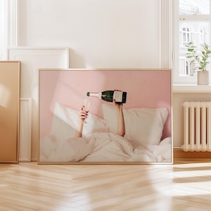 May include: A framed print depicts a person pouring champagne into a glass while in bed. The image features a green champagne bottle, a clear glass, and white bedding against a pink backdrop. The person's hands are visible.