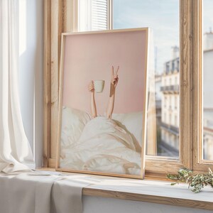 May include: A framed print featuring a person's arms reaching up from under a white blanket. One hand holds a white mug, the other makes a peace sign. The background is a soft pink, and the frame is a light wood color.