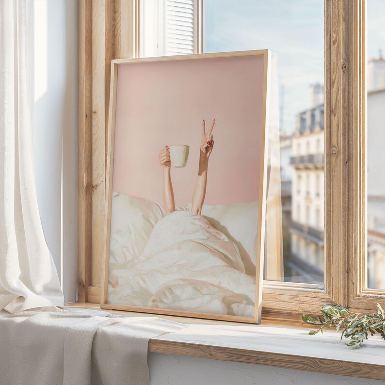 May include: A framed print featuring a person's arms reaching up from under a white blanket. One hand holds a white mug, the other makes a peace sign. The background is a soft pink, and the frame is a light wood color.