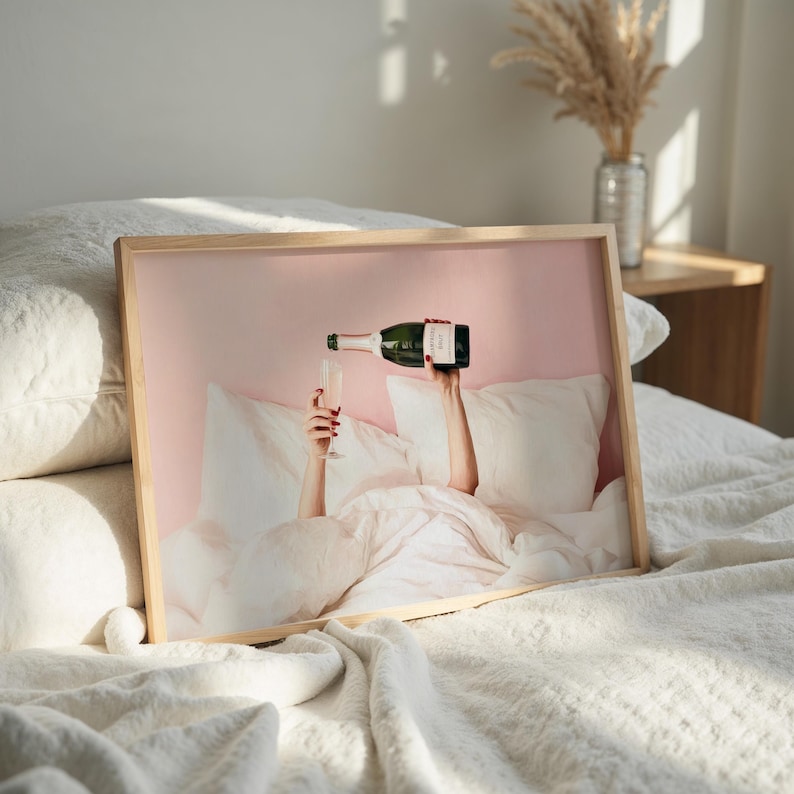 May include: A framed print featuring a person pouring champagne into a glass while in bed. The image has a pink background and a light wooden frame. The person's hands are visible, holding the champagne bottle and glass.