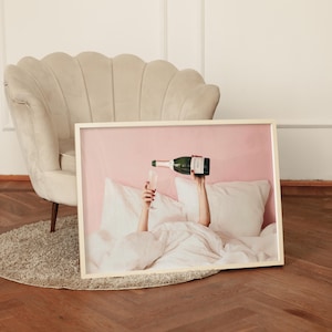 Puede incluir: Una impresión enmarcada muestra a una persona sirviendo champán en una copa en la cama. La imagen tiene un fondo rosa y presenta almohadas y ropa de cama blancas. La botella de champán es verde con una etiqueta negra.