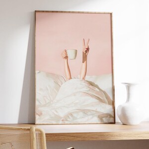 May include: A framed art print depicts a person in bed, holding a white mug and making a peace sign. The background is a soft pink, and the bed linens are white. The artwork is displayed on a wooden shelf with a white vase.
