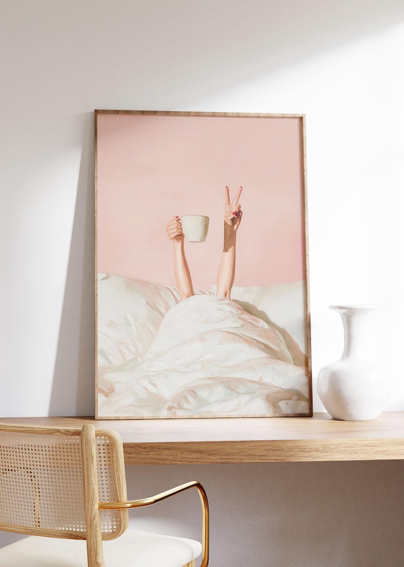 May include: A framed art print depicts a person in bed, holding a white mug and making a peace sign. The background is a soft pink, and the bed linens are white. The artwork is displayed on a wooden shelf with a white vase.