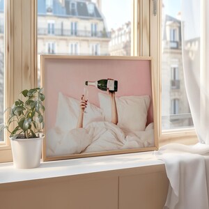 May include: A framed print featuring a champagne bottle pouring into a glass, with arms reaching up from under white bedding. The background is a soft pink, and the image is set in a light wood frame. The scene is set by a window.