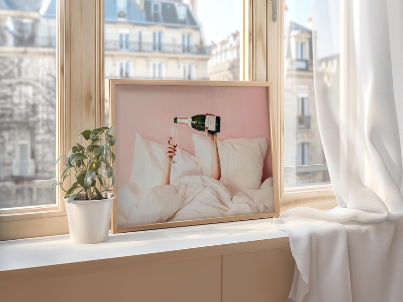May include: A framed print featuring a champagne bottle pouring into a glass, with arms reaching up from under white bedding. The background is a soft pink, and the image is set in a light wood frame. The scene is set by a window.