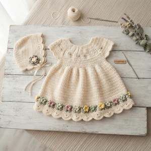 Peut inclure: Une robe en crochet couleur crème à manches courtes avec un bonnet assorti. La robe présente un ourlet festonné orné de petites applications florales multicolores en rose, jaune et vert. Une étiquette en bois marquée HANDMADE est incluse.