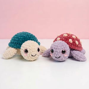 Peut inclure: Deux tortues en crochet en peluche faites de fil doux. La tortue de gauche a une carapace sarcelle et une tête et des membres couleur crème. La tortue de droite a une carapace bordeaux avec des taches crème et une tête et des membres lavande.