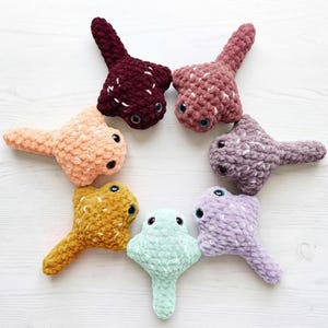 Peut inclure: Sept jouets en peluche en forme de raie disposés en cercle, chacun fabriqué à partir de fil épais et doux dans des couleurs distinctes dont bordeaux, vieux rose, pêche, jaune moutarde, vert menthe, lavande et violet, avec des yeux en plastique.