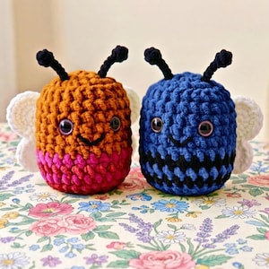 Peut inclure: Deux peluches en crochet en forme d'abeille avec de grands yeux sombres et des antennes. L'abeille de gauche est orange et rose, tandis que celle de droite est bleue et noire. Elles ont des ailes blanches en crochet et une texture en laine.