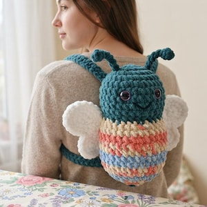 Peut inclure: Un sac à dos en crochet en forme d'abeille avec une tête bleu sarcelle foncé, deux antennes et des yeux violets. Le corps présente des rayures horizontales pêche, crème et bleu clair. Deux ailes blanches en crochet sont fixées sur les côtés.