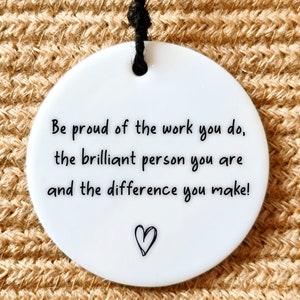 May include: A white round ceramic ornament with a black string for hanging. The ornament features a black handwritten message that reads: "Be proud of the work you do, the brilliant person you are and the difference you make!" A small black heart is drawn at the bottom of the message.