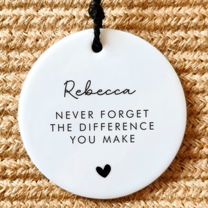 May include: White round ceramic ornament with black text that reads "Rebecca Never Forget The Difference You Make" and a small black heart.