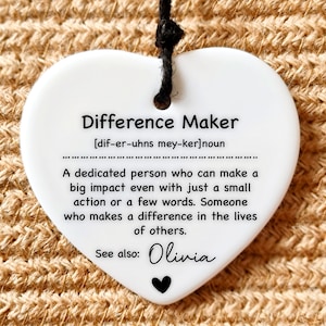 May include: White heart-shaped ceramic ornament with a black string for hanging. The ornament has the text "Difference Maker" with a definition and the name "Olivia" written below.