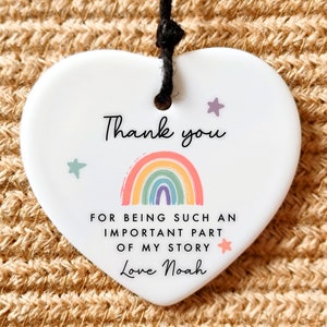Puede incluir: Adorno de cer&aacute;mica blanca en forma de coraz&oacute;n con un arco&iacute;ris y estrellas. El adorno dice "Gracias por ser una parte tan importante de mi historia. Con amor, Noah."