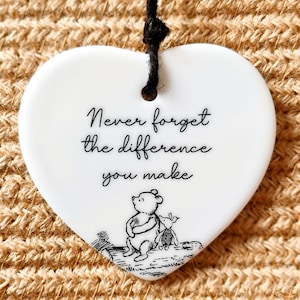 May include: White ceramic heart-shaped ornament with a black string for hanging. The ornament features a black and white illustration of Winnie the Pooh and Piglet. The text "Never forget the difference you make" is printed in black on the ornament.