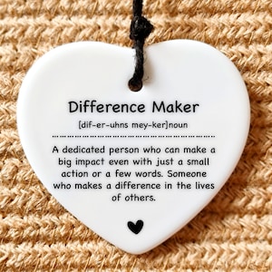 May include: White ceramic heart-shaped ornament with a black string for hanging. The ornament has the text "Difference Maker" with a definition: "[dif-er-uhns mey-ker]noun A dedicated person who can make a big impact even with just a small action or a few words. Someone who makes a difference in the lives of others."