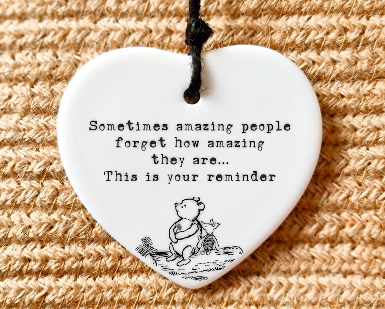 May include: White heart-shaped ceramic ornament with black text: "Sometimes amazing people forget how amazing they are... This is your reminder." Features a Winnie the Pooh illustration. Includes a black string for hanging.