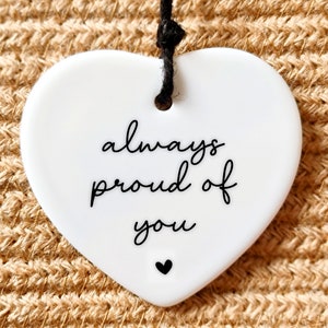 May include: White ceramic heart-shaped ornament with black string for hanging. The ornament has the text "always proud of you" written in black cursive font with a small black heart at the bottom.