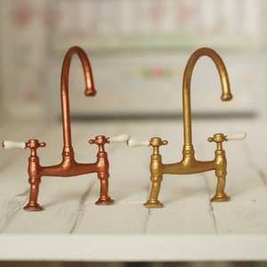 Op de afbeelding: Twee miniatuur keukenkraan, een koperen en een gouden, met witte porseleinen handvatten.