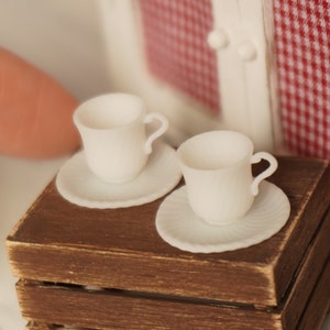 Könnte beinhalten: Zwei weiße Miniatur-Teetassen und Untertassen auf einer braunen Holzkiste. Die Tassen und Untertassen sind aus Keramik und haben ein schlichtes Design.