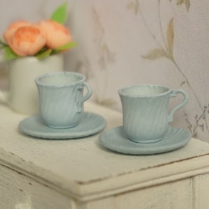 Könnte beinhalten: Zwei hellblaue Miniatur-Teetassen und Untertassen stehen auf einer weißen Oberfläche. Die Teetassen haben ein strukturiertes, geriffeltes Design und kleine Henkel. Im Hintergrund befindet sich ein weißer Becher mit pfirsichfarbenen Blumen.
